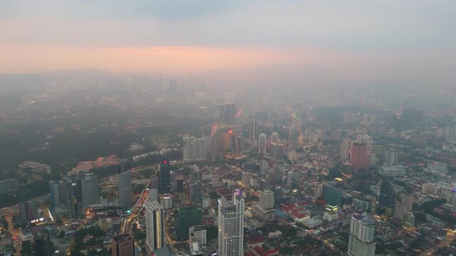 Aerial view across Kuala Lumpur with KL Tower centred and Merdeka 118 distant. The movement tracks from gardens to towers while haze softens mountains, traffic sparkles and the skyline breathes depth