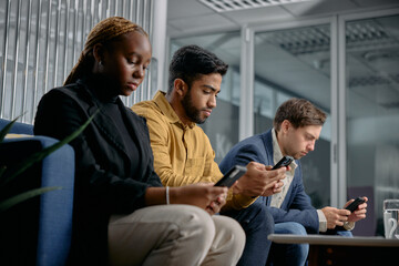 Three multiracial young adults in businesswear sitting on sofa and typing on mobile phones in office