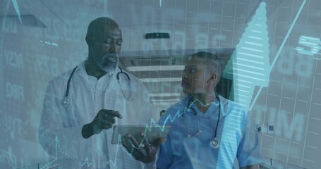 Wearing lab coat doctor showing tablet to scrub clad colleague in hospital hall, with data overlays