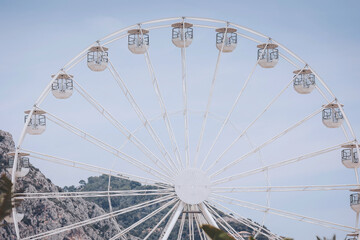 Fototapeta premium Majestic ferris wheel gracing the skyline against a backdrop of serene mountains and a clear blue sky during a sunny afternoon