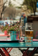Cozy turquoise metal table with red legs holding a glass of water, a beer glass, and a glass water carafe, with blurred bucket of pink flowers and city atmosphere in the background.