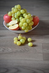 A fresh assortment of green grapes and red apples, one apple sliced ​​to show its core, in a white bowl on a wooden table. A few grapes are scattered on the table, highlighting the natural feel.