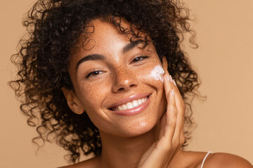 Young woman of African descent with curly black hair, light brown skin and freckles applying white cream to her face with fingers and looking at camera smiling. Healthy glowing skin. Generative AI