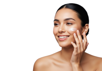 Young mixed-race woman, dark hair in bun, bare shoulders, happily applying cream to cheek on transparent studio background with copy space, self-care and beauty concept