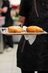 Chef holding tray with two golden fried croquettes in commercial kitchen. Freshly cooked street food served in professional setting. Culinary preparation concept.
