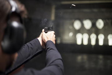 Caucasian man practicing shooting at indoor firing range, holding handgun with both hands, wearing protective earmuffs, bullet visible in midair, targets blurred in background © DragonImages
