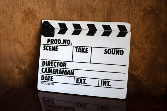Classic film clapperboard resting on a dark, reflective surface against a warm wooden background. Symbolizes filmmaking, production, and cinema.