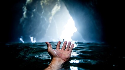 A human hand emerges from dark, rippling water in a mysterious cave, reaching towards a brilliant,...