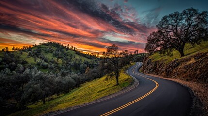 Fototapeta premium A winding road curves through lush green hills under a vibrant sunset sky filled with orange and purple hues.