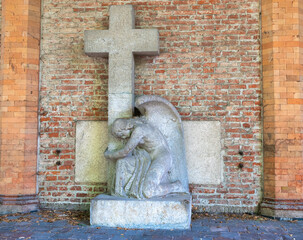 ein Engel kniet vor einem Grabstein mit Kreuz vor einer Ziegelmauer Alter Südfriedhof München