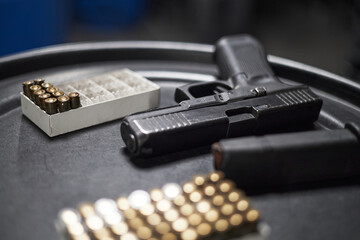 Handgun resting on round black surface with loaded magazine nearby, boxes of ammunition arranged in foreground and background, focus on firearm and bullets, no people visible