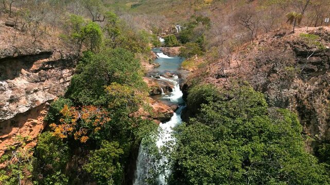 A imagem a&eacute;rea destaca uma cachoeira caudalosa despencando entre rochas e vegeta&ccedil;&atilde;o densa. A &aacute;gua branca contrasta intensamente com o verde vibrante das &aacute;rvores e o tom terroso das pedras ao redor.