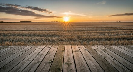 Golden sunset over a vast harvested field with a rustic wooden deck in the foreground, offering a serene rural backdrop with warm light and sun rays.