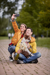caucasian woman taking selfie with her boyfriend and his dog in a lap, sitting on a path in park