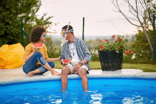 Silly couple sitting by the pool in swimming goggles eating watermelon fruit and talking.