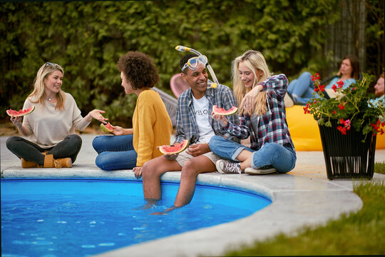 Lovely young adults sitting by the pool outdoors enjoying time together and eating watermelon fruit.