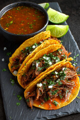 Juicy beef Birria tacos served with bowl Consommé soup sauce, lime on dark black slate platter close up detail.