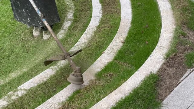 Worker trimming grass edges along stone steps and pavement with a string trimmer. Lawn care maintenance highlighting precise landscaping and clean borders between grass and concrete.