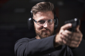 Portrait of Caucasian young adult man wearing protective glasses and earmuffs aiming handgun at shooting range, focusing on target with intense expression, hands extended forward