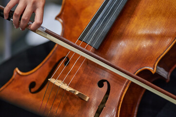 A close-up of a musician&rsquo;s hand moving a bow across the strings of a cello, highlighting the instrument&rsquo;s smooth, rich wooden surface and the precision of the musician