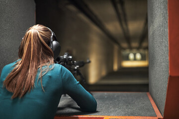 Caucasian young adult woman wearing protective headphones aiming rifle at shooting range targets, seen from behind with long hair tied back, focusing on precision practice
