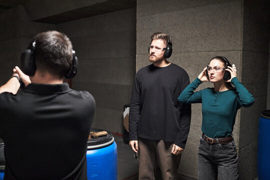Caucasian young adult man and Caucasian young adult woman standing at indoor shooting range wearing protective headphones and glasses while instructor demonstrating firearm handling