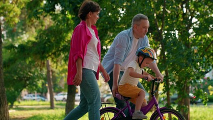 Fototapeta premium Child cyclist with parents in city park. Family weekend, father teaches son to ride bike in park. Happy family, little child in helmet with father and mother learning to ride bike outdoors in summer
