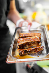 Close-up of glazed barbecue ribs served on a metal tray, held by a person in a kitchen setting. Juicy, saucy meat with rich texture, ideal for food photography and culinary themes.