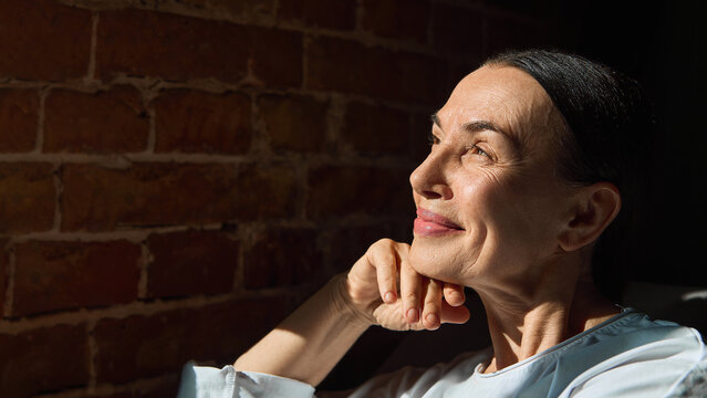 Mature woman in white shirt smiling towards sunlight near brick wall. Concept of skincare, aging wellness, lifestyle positivity and natural grace with hopeful emotion. - Powered by Adobe