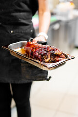 Cooked pork shoulder with crispy crust served in metal tray, held by chef in black uniform. Juicy roast meat in kitchen setting, ideal for culinary, restaurant, or food service themes
