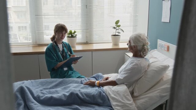 Zoom in view of young female medical worker with clipboard asking senior woman questions and taking notes while visiting her in hospital room, patient lying in bed with nasal cannula