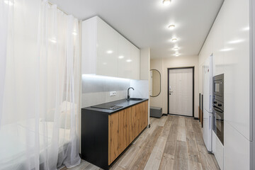 well-lit, modern apartment featuring a compact kitchen with sleek white cabinets, a black countertop, and wooden accents. Sheer curtains separate the kitchen from the living area