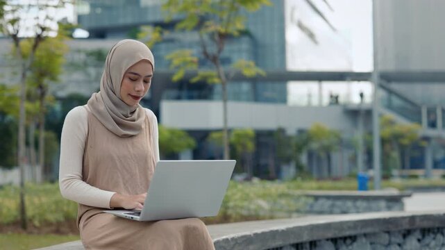 Portrait of positive Malaysian woman wearing hijab types on laptop sits on low wall parapet. Young Muslin female focuses on work tasks uses notebook computer in downtown district