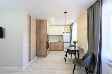 modern, compact kitchen with light wood cabinets, white upper cabinets, a small dining table, and gray curtains. Overall, the space is bright and airy
