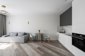 Modern apartment interior featuring a gray sofa, wooden floor, dining table with two chairs, and a white kitchen with black appliances