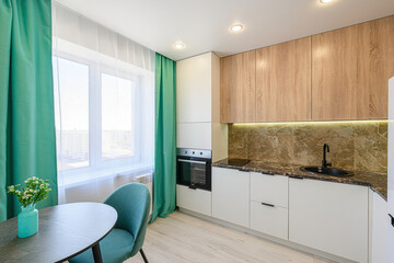 Bright kitchen with wooden cabinets, marble countertops, and a table near the window with green curtains. A simple, modern design