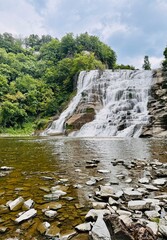 Obraz premium Majestic waterfall cascading over layered rock formations into a calm pool, surrounded by greenery and cliffs.