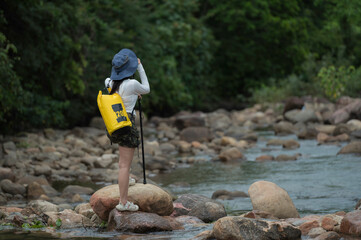 Young woman carrying a yellow backpack and camera is hiking through nature, carefully crossing a rocky stream in the forest. Outdoor adventure and eco-travel lifestyle concept.	