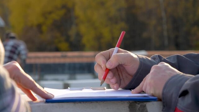 Engineer drawing sketch with pencil on construction site, receiving instructions from a colleague pointing finger on white sheet of paper attached to a clipboard. Close up hands builders workers