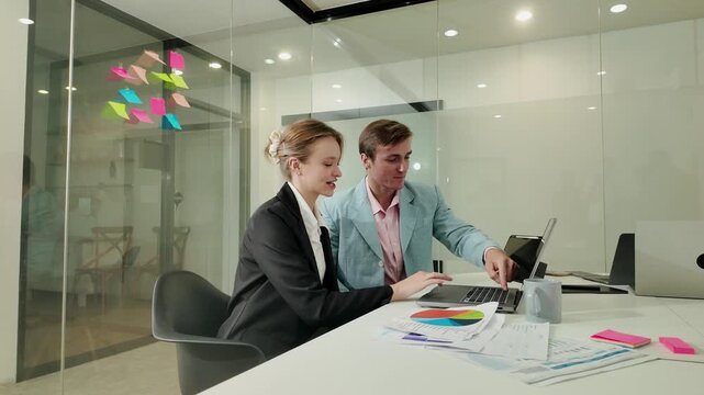 Two Business People Collaborating: A man and woman in business attire collaborate intently on a laptop in a modern office, surrounded by colorful sticky notes and papers on a conference table.