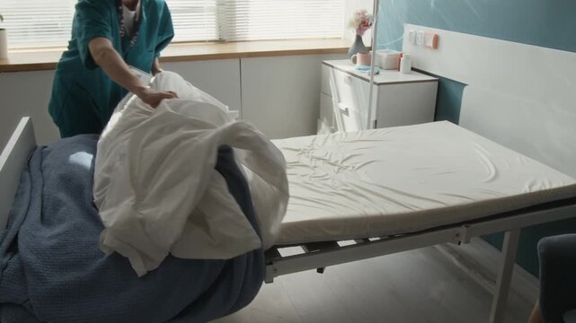 Zoom in shot of young nurse wearing scrubs removing pillow, sheet and blankets off patients bed after their discharge from hospital