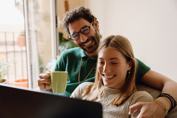 A man is holding a cup and hugging a woman who is holding a laptop, which they are looking at and smiling at