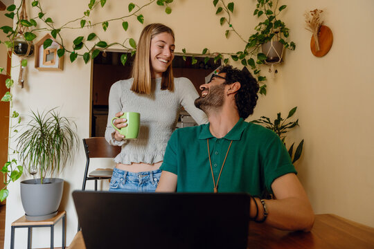 A woman is holding a cup and standing next to a man who is sitting at a table in front of a laptop while they are smiling