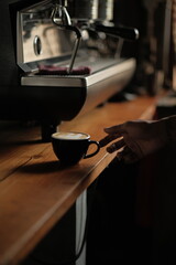 A barista’s hand reaches for a hot coffee cup on a wooden counter in a cozy café. A modern coffee machine is visible, highlighting the moment of serving a fresh brewed latte .