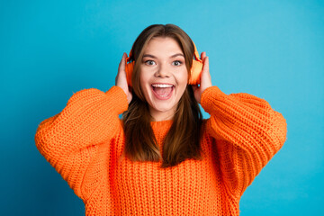 Smiling young woman in vibrant orange knit sweater enjoying music with headphones against a cheerful blue background
