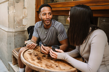 A male athlete is holding a phone and talking to a female athlete who is sitting next to him at a table and holding a cup