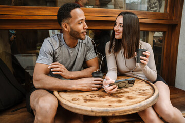 Male and female athlete listening in earphones and sitting at a table while holding cups and she...