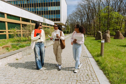 A group of three female students walk and talk while holding folders, notebooks, and a cup