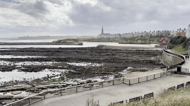 Slow pan shot across a rocky harbour bay and sandy beach at low tide, overlooked by properties facing the sea from the local town.