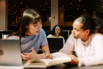 A female student smiles and looks at a laptop while two female students talk in front of her while they sit at desks